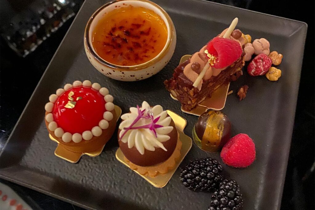 An elegant assortment of miniature desserts on a dark plate, featuring a crème brûlée, a raspberry chocolate tart, a red glazed mousse, and fresh blackberries.