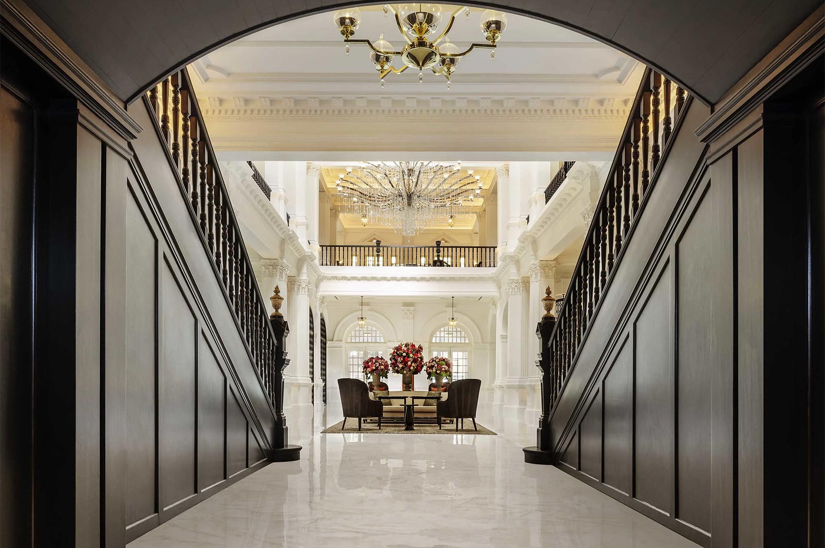 Symmetrical view of a grand hotel lobby featuring dark wood staircases, white marble floors, and a massive crystal chandelier.