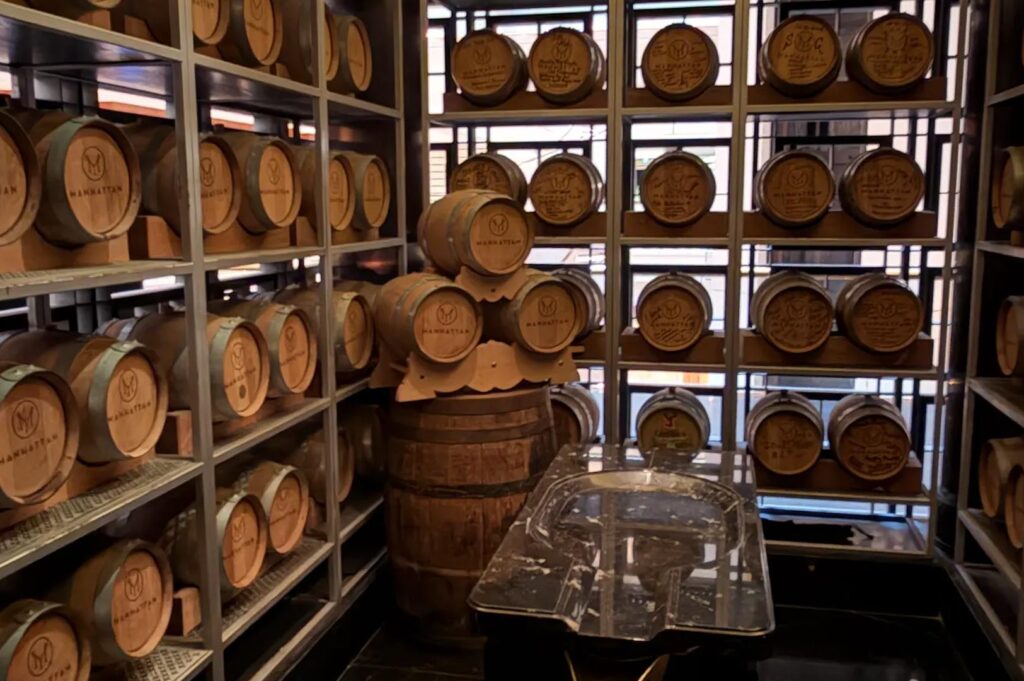 A specialized rickhouse room featuring rows of small oak barrels on metal racks used for in-house cocktail aging.