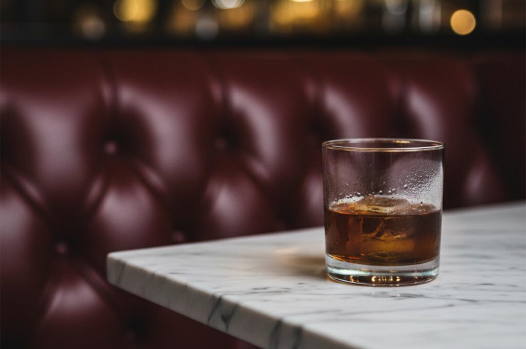 A close-up of an Old Fashioned cocktail with a large ice cube on a marble table against a red leather booth.