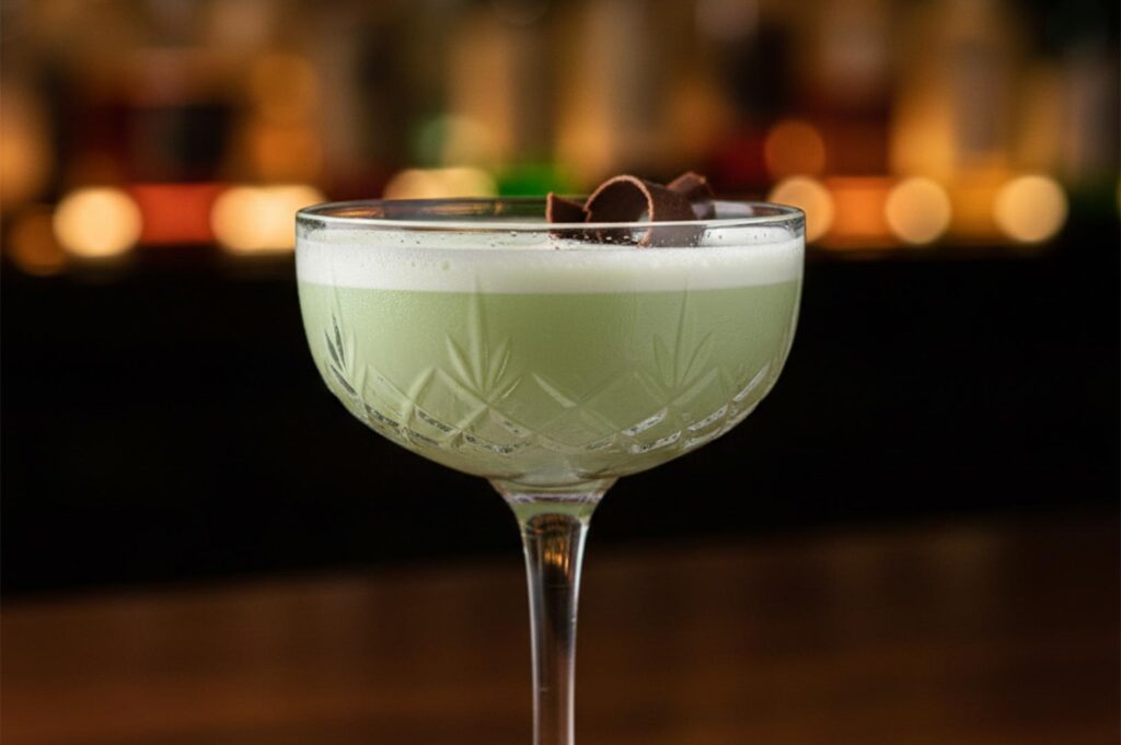 A pale green creamy cocktail served in a crystal coupe glass, topped with a frothy foam layer and a delicate chocolate curl garnish against a blurred bar background.