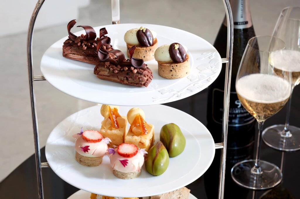 Close-up of a three-tier afternoon tea stand featuring chocolate eclairs, fruit tarts, and macarons next to glasses of champagne.