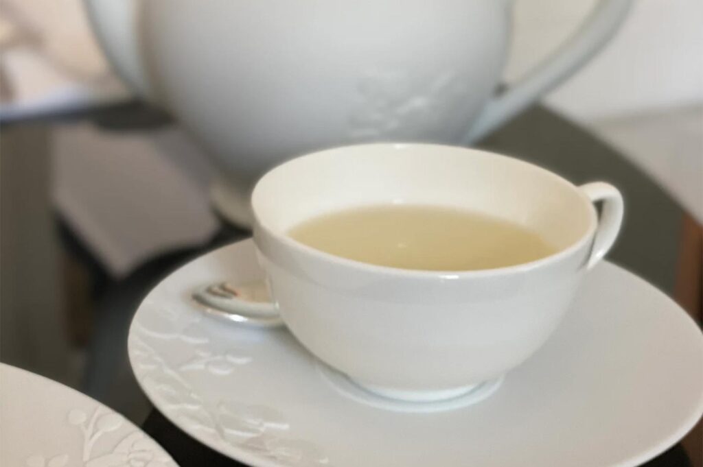Close-up of a delicate white porcelain teacup filled with light herbal tea, with a matching teapot in a soft-focus background.