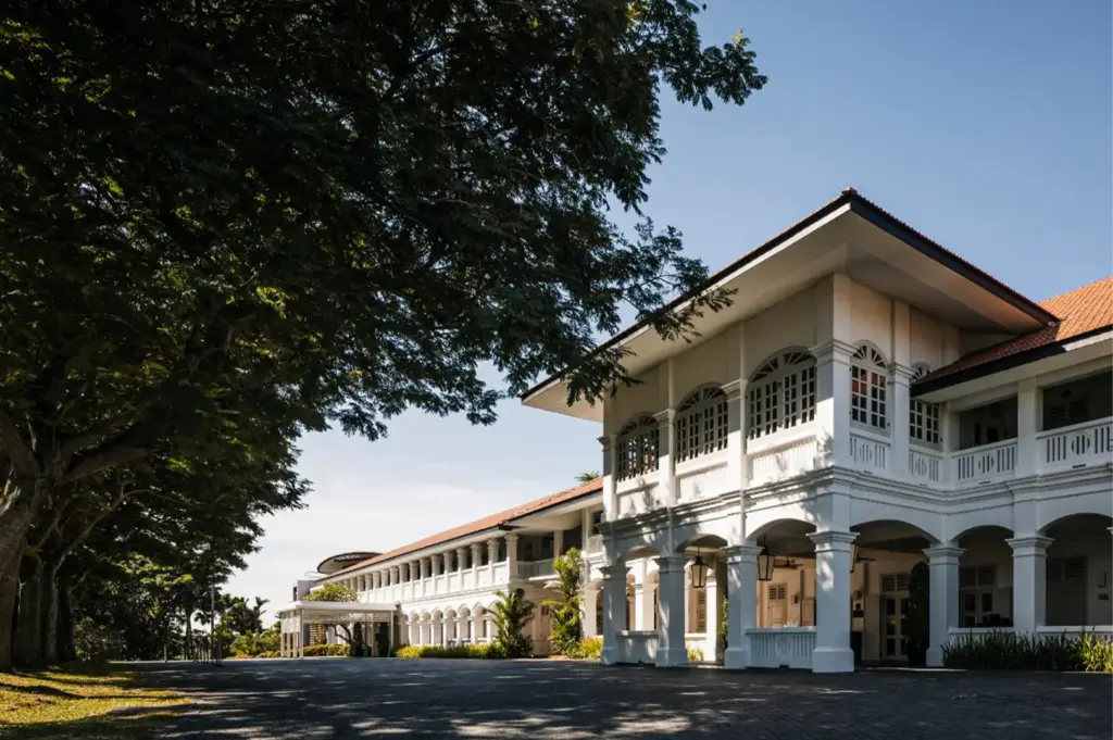 An eye-level, wide shot captures the majestic facade of a colonial-style luxury estate, representing the file "1-Best Singapore Hotels Culinary Journeys.webp". The pristine, two-story white building features a prominent grand entrance on the right supported by sturdy pillars, classic arched windows, and an upper-level balcony, all beneath a warm terracotta-tiled roof. A long wing of matching arched corridors extends into the background, bordering a wide, dark asphalt driveway. On the left, a row of mature, leafy green trees casts a deep canopy of shade over the grassy roadside, contrasting beautifully with the bright, clear blue sky overhead.