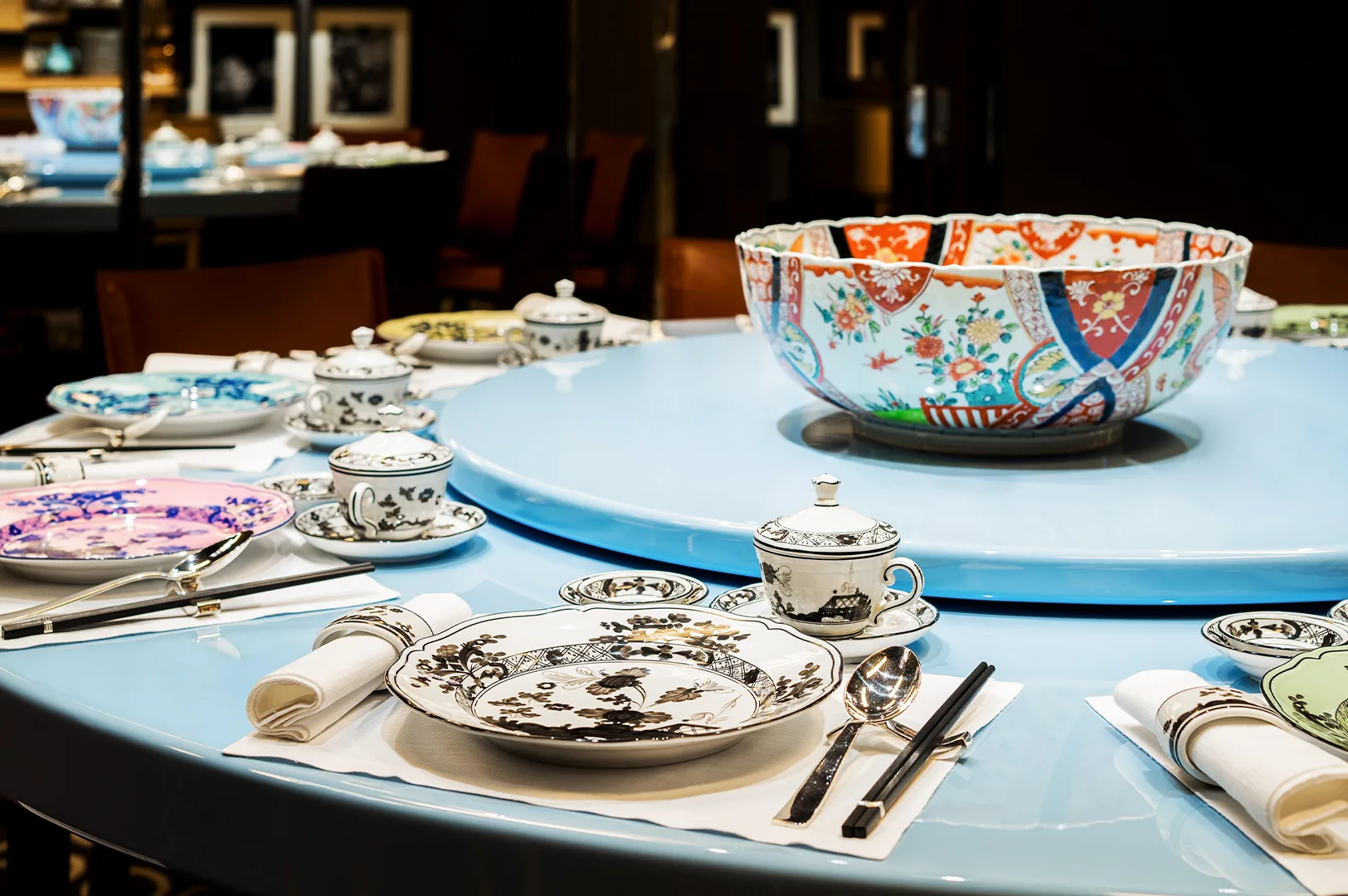 Elegant dining setup with colorful patterned plates, teacups, and napkins on a light blue round table. A large decorative bowl is at the center.