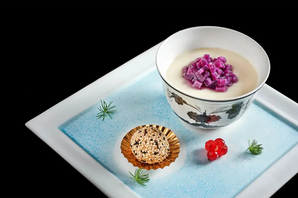 A gourmet Cantonese dessert set featuring a bowl of creamy almond pudding topped with diced purple yam, served alongside a toasted sesame ball.