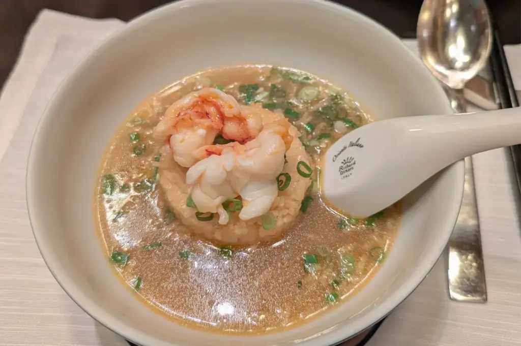 A bowl with a rice dish topped with shrimp and garnished with chopped green onions in clear broth. A ceramic spoon rests on the dish.