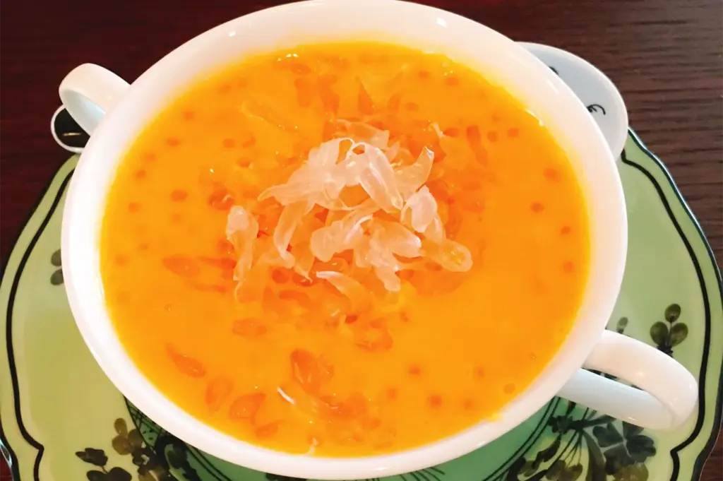 A vibrant bowl of chilled mango sago cream topped with fresh pomelo segments and translucent sago pearls, served in a decorative white porcelain cup.