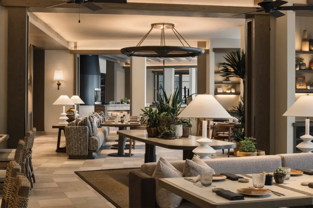 An elegant, contemporary lounge and dining area with warm lighting, neutral-toned furniture, and lush indoor potted plants.