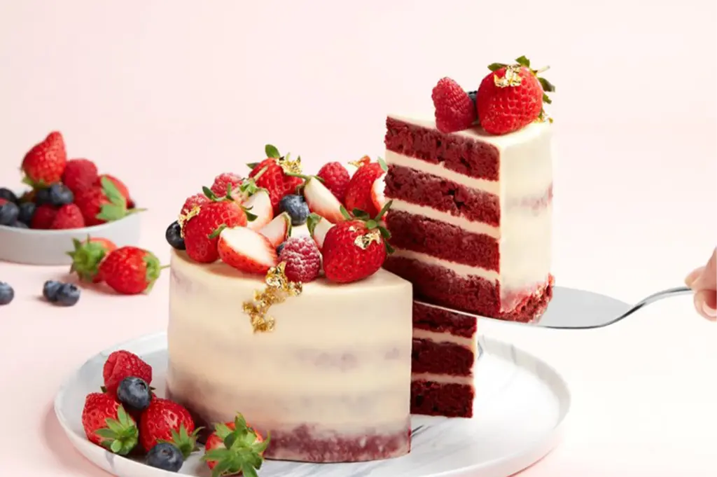 A slice of rich red velvet cake with white frosting is being lifted. The cake is topped with fresh strawberries, blueberries, and gold leaf, creating an elegant, celebratory tone.