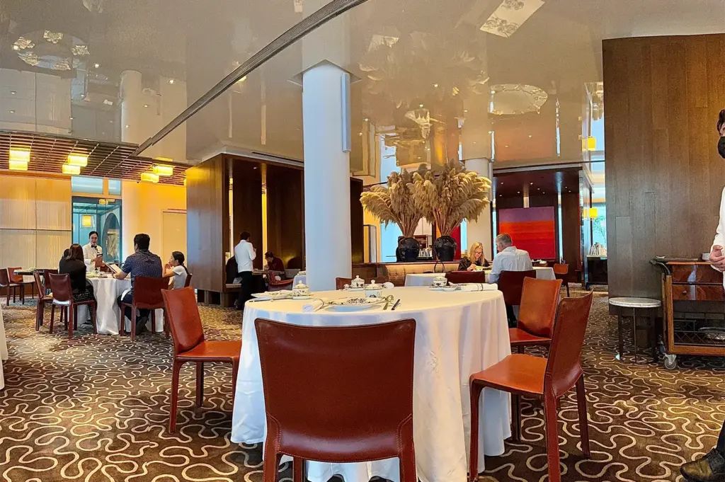 Elegant restaurant interior with round tables draped in white cloths, brown chairs, and a decorative centerpiece. Diners are engaged in conversation, creating a warm, inviting atmosphere.