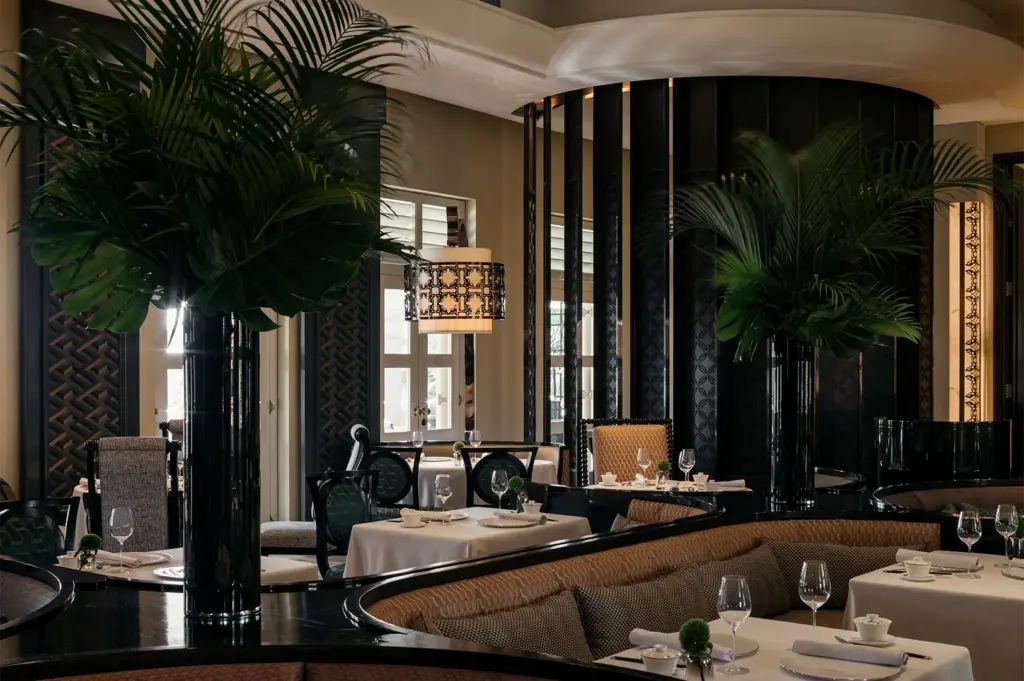 A sophisticated restaurant interior featuring curved booths, white tablecloths, and large decorative indoor palm trees in black pillars.