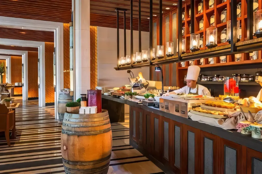 A professional chef working behind a sleek, modern buffet counter with warm wood accents and hanging light fixtures.