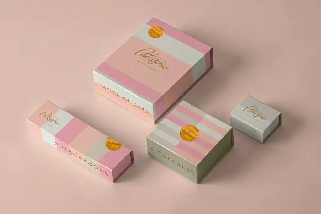 Pastel-colored pastry boxes on a soft pink background. The boxes, labeled for cake, macaroons, and cupcakes, feature elegant gold lettering and seals.