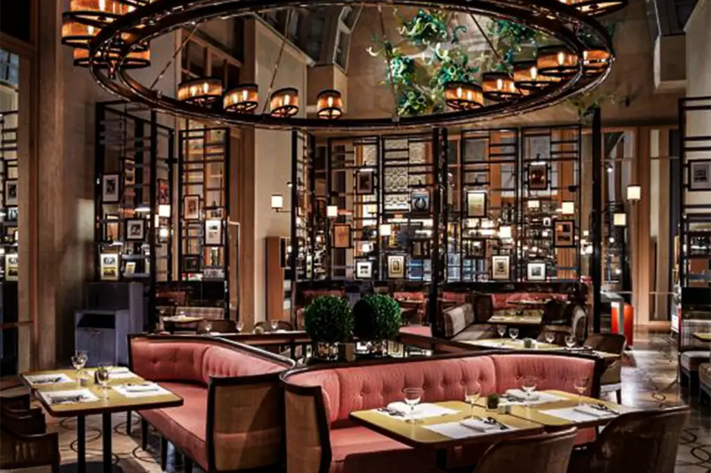 An eye-level, medium shot captures the lavish and meticulously designed interior of an upscale hotel restaurant, representing the file "6-Best Singapore Hotels Culinary Journeys.webp". In the foreground, curved, tufted pink banquette sofas sit back-to-back, paired with neatly set dining tables featuring crisp white placemats, wine glasses, and polished silverware under the soft glow of a massive, circular wrought-iron chandelier. Separating the intimate dining booths from the rest of the room are tall, intricate black metal grid partitions decorated with small, framed pictures and built-in lights. In the background, a high glass-atrium ceiling reveals a vibrant, green-and-blue abstract glass sculpture hanging overhead, adding an artistic, grand touch to the warm and sophisticated ambiance.
