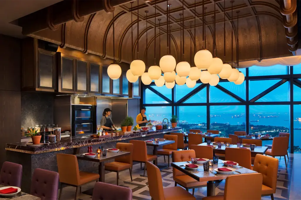An open kitchen and dining area with orange chairs, globe pendant lights, and floor-to-ceiling windows overlooking the ocean at dusk.