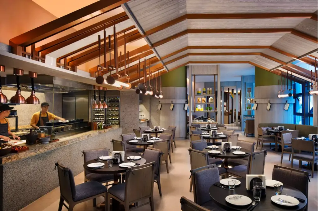 An eye-level, medium shot captures the sleek and modern interior of a bustling hotel restaurant, representing the file "9-Best Singapore Hotels Culinary Journeys.webp". On the left, an open, glass-enclosed kitchen features stone counters where chefs in yellow shirts and black aprons prepare food under warm, copper heat lamps and modern hanging light fixtures. The main dining area is filled with neatly arranged round wooden tables paired with comfortable, fabric-upholstered grey chairs, each table set with white plates, glasses, and cutlery. The sophisticated space boasts a unique, angled ceiling lined with light wood panels and copper beams, while the brightly lit background reveals additional dining sections and decorative shelves, creating an inviting and upscale culinary environment.