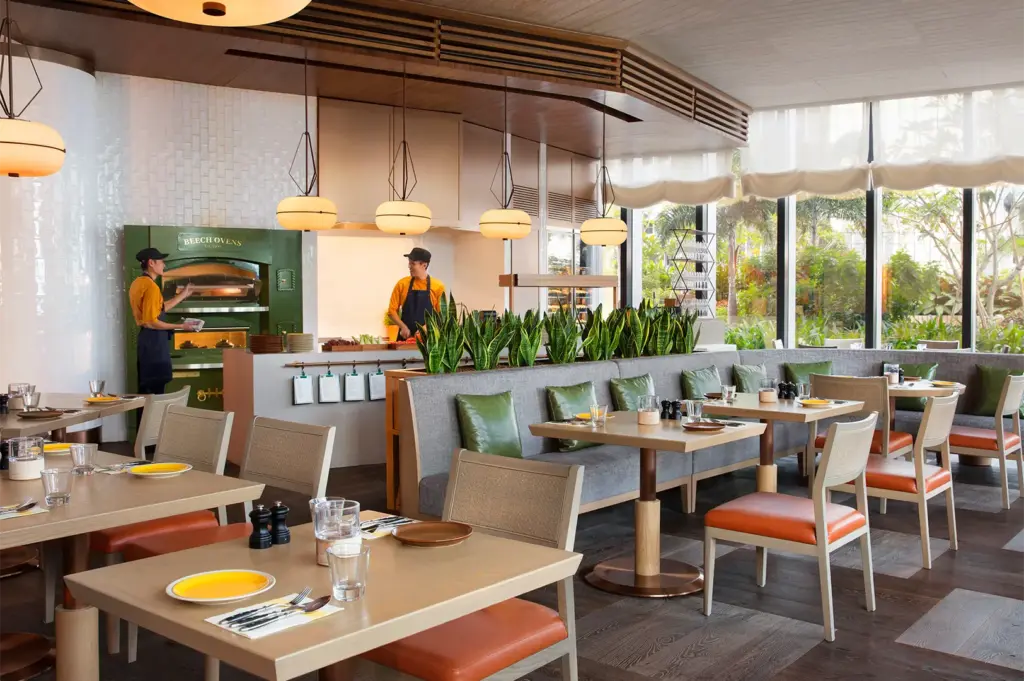 A bright, modern bistro featuring a green Beech Ovens wood-fired oven and chefs preparing food behind a counter lined with snake plants.
