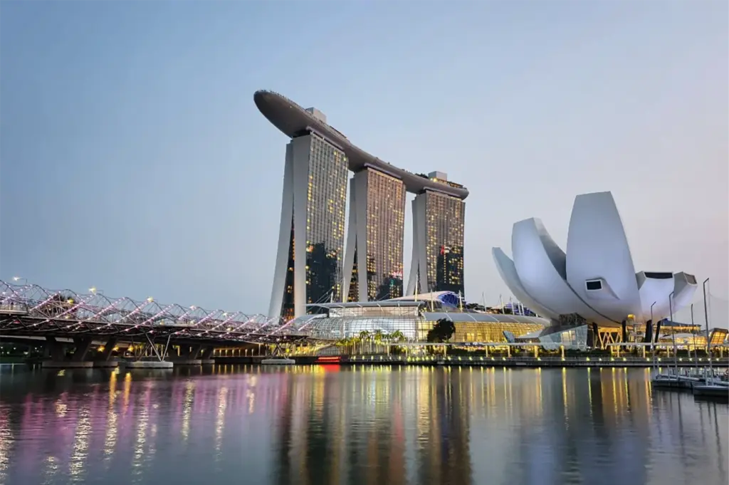 An eye-level, wide shot captures the iconic, illuminated skyline of Singapore's waterfront district at dusk, representing the file "1-Where to Dine at Marina Bay Sands.webp". Centered in the background stands the massive Marina Bay Sands hotel, with its three distinct skyscraper towers connected at the top by a vast, boat-shaped skypark terrace. To the right, the striking white, lotus-inspired structure of the ArtScience Museum sits prominently at the water's edge, while the Helix Bridge, glowing with pink and purple lights, curves across the left side of the frame. In the foreground, the calm, glassy surface of the bay perfectly mirrors the vibrant lights of the modern architectural landmarks under a clear, transitioning evening sky.
