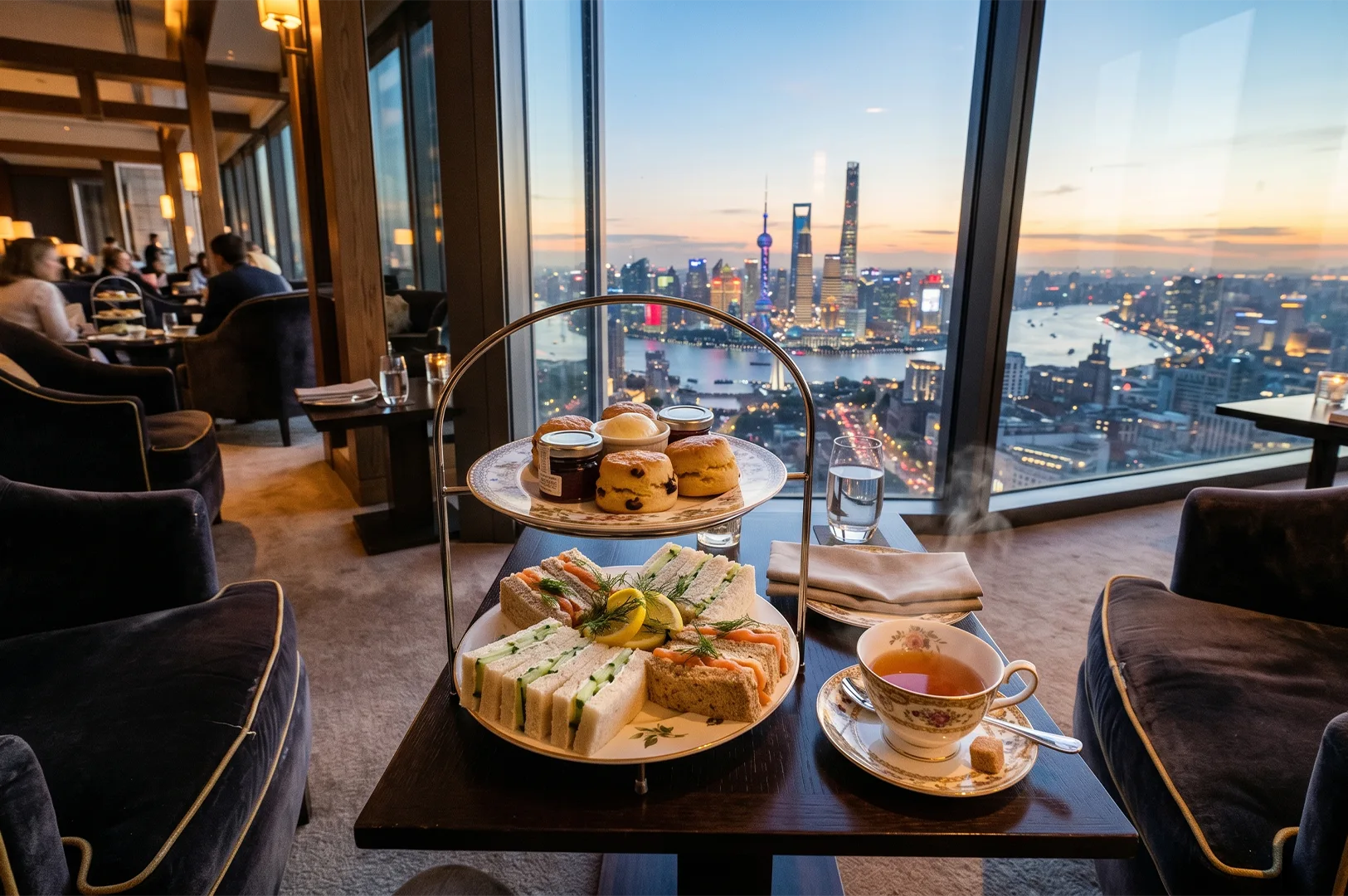 A luxury afternoon tea set with tiered stands of sandwiches and scones on a wooden table, overlooking the Shanghai Lujiazui skyline and Oriental Pearl Tower at sunset.