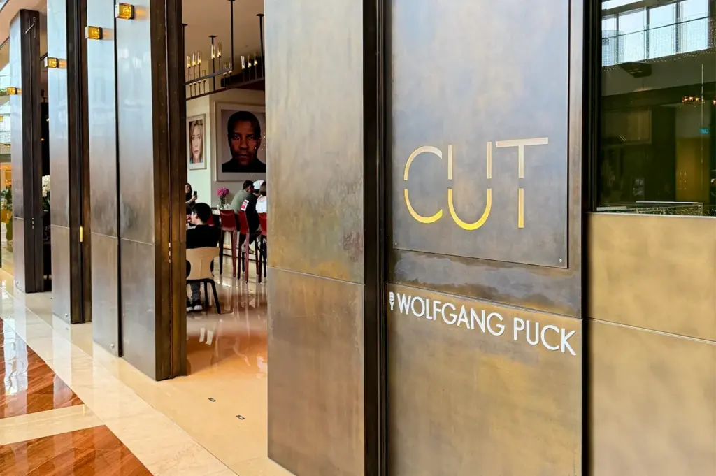 The modern brass-clad entrance of CUT by Wolfgang Puck restaurant, featuring a view into the stylish dining room with celebrity portraits.