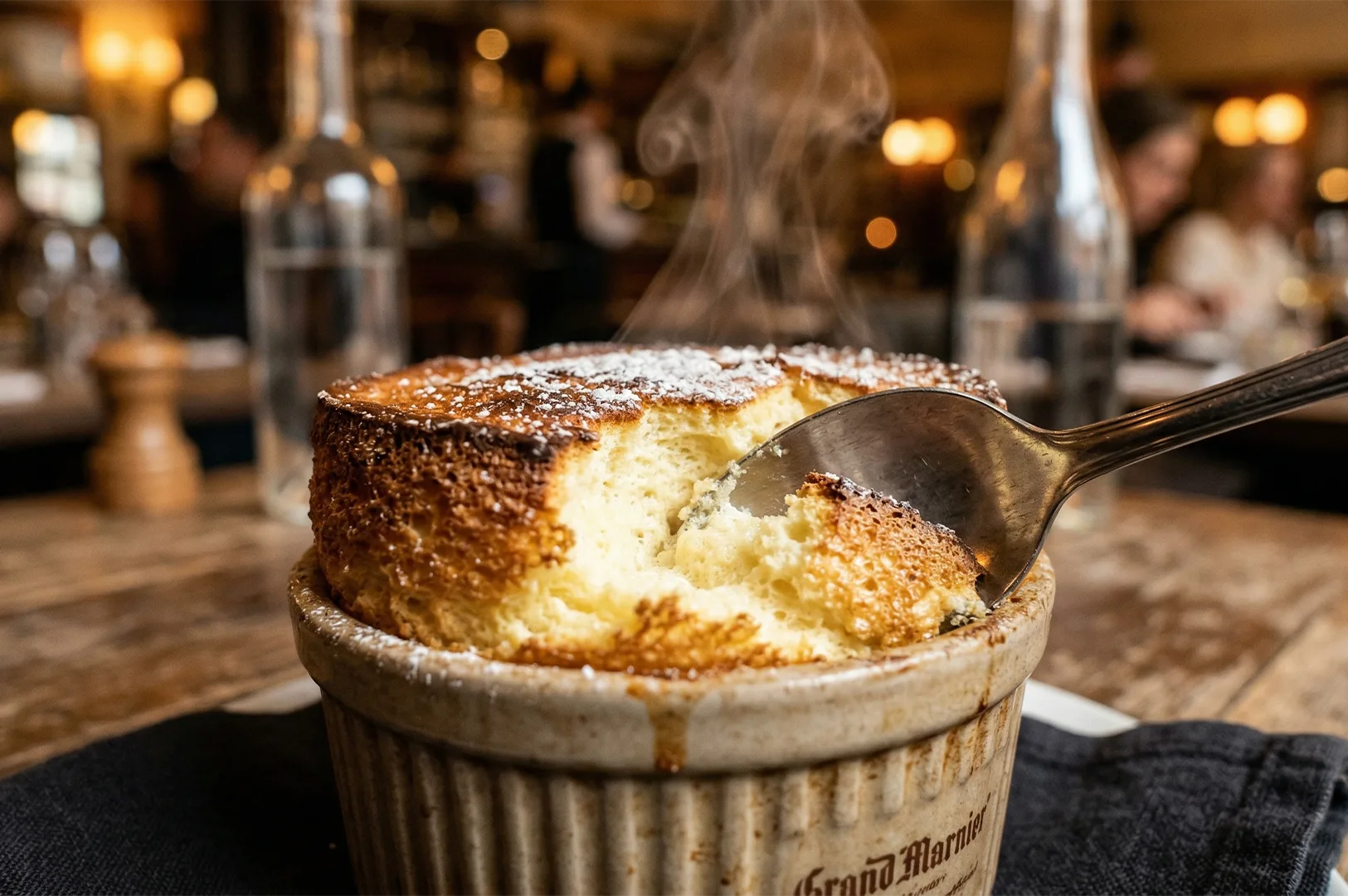A steaming Grand Marnier soufflé with a golden-brown crust, dusted with powdered sugar, is freshly scooped by a spoon in a cozy restaurant setting.