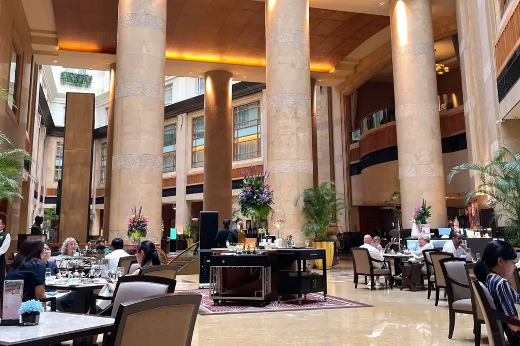 A wide shot of a sunlit, high-ceilinged hotel atrium with massive stone pillars, palm plants, and guests enjoying afternoon tea at The Courtyard.