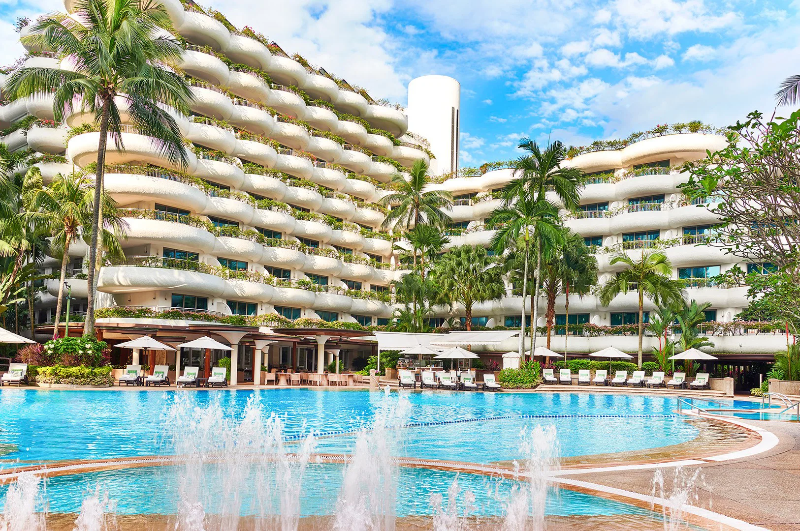 A luxurious tropical resort featuring a large blue swimming pool with fountains, surrounded by white curved balconies with lush greenery and palm trees under a blue sky.