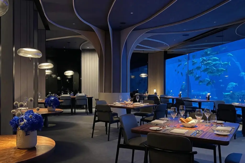 Sophisticated underwater restaurant with dim lighting, tables set elegantly with glassware and flowers. Large aquarium window reveals swimming fish, creating a serene ambiance.