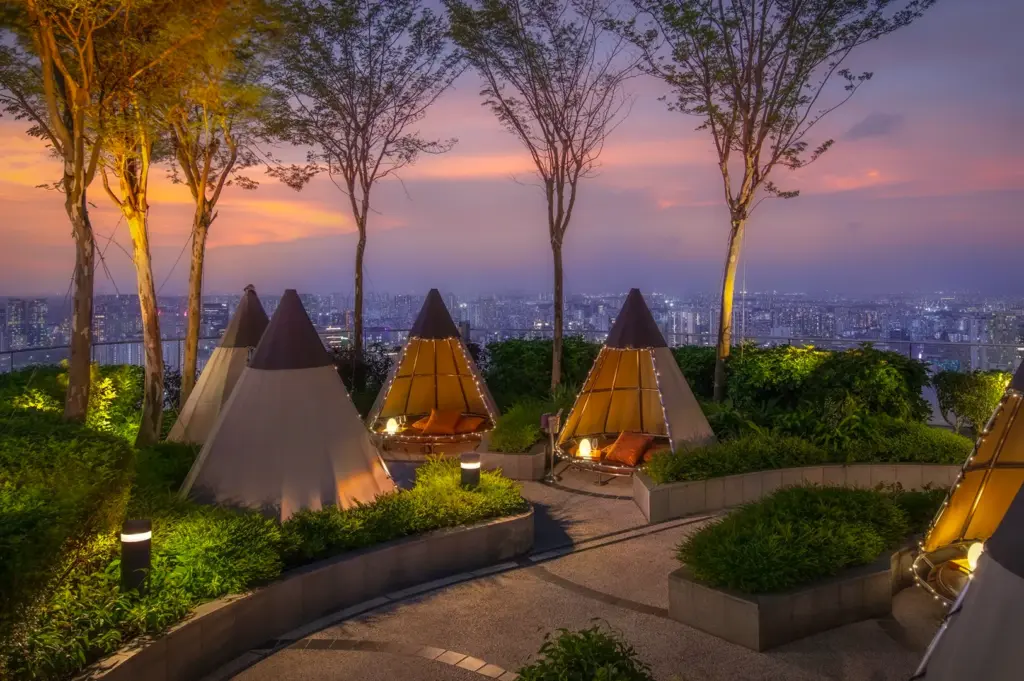 Stunning panoramic rooftop view from Mr. Stork at Andaz Singapore during sunset, featuring cozy illuminated teepee huts nestled among lush greenery with the city's sparkling skyline in the background.