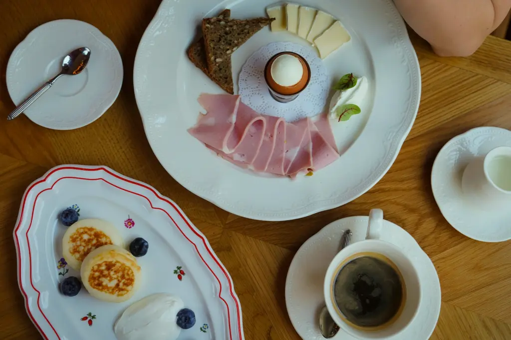 Luxurious breakfast spread on a wooden table featuring a soft-boiled-egg, sliced ham, brie cheese, seeded bread, Syrniki pancakes with blueberries, cream, and a fresh cup of black coffee.