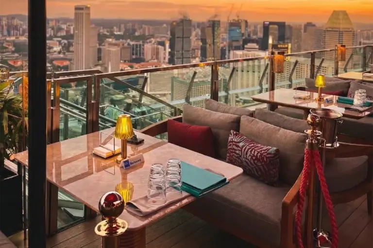 Rooftop restaurant at sunset, overlooking a city skyline. Cozy seating with plush cushions, elegant table settings, and a glass railing enhancing the view.