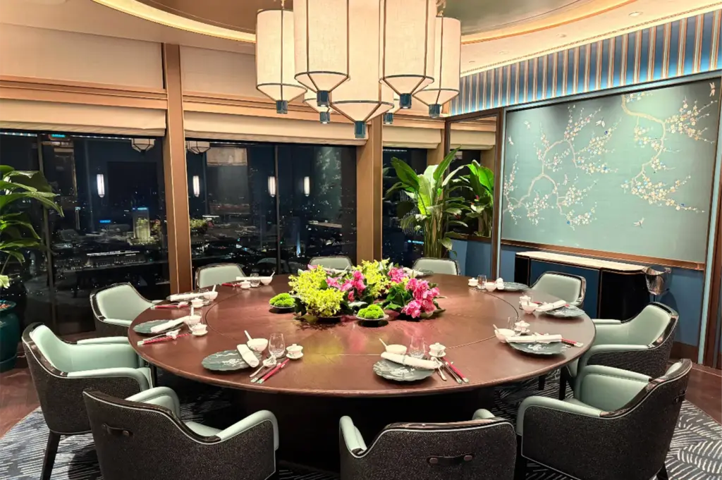 An eye-level, medium shot captures a luxurious, high-end private dining room set up for a large group, representing the file "10-Where to Dine at Marina Bay Sands.webp". A massive, round dark wood dining table dominates the room, encircled by plush, high-backed armchairs upholstered in a striking mint green and dark textured grey fabric. The table features an elegant centerpiece arrangement of vibrant pink orchids and bright green-yellow hydrangeas, surrounded by meticulously set individual place settings with patterned teal plates, white porcelain bowls, cups, and chopsticks. Hanging directly above the table is a grand, multi-tiered white fabric lantern chandelier that casts a soft glow across the room. In the background, expansive floor-to-ceiling windows look out onto a dark city skyline at night, while the interior walls are accented with a beautiful tiffany blue panels, including a large framed artwork of white floral branches, completing the sophisticated and serene ambiance.