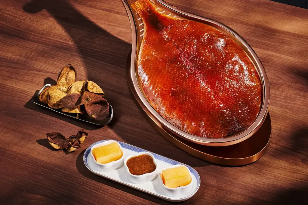 A high-angle, close-up shot captures a meticulously presented culinary dish, representing the file "11-Where to Dine at Marina Bay Sands.webp". Arranged on a rich, dark brown wooden surface, the center attraction is a large piece of roasted meat with glistening, crispy, golden-brown skin, shaped and displayed elegantly inside a copper, lute-shaped platter. To the left sits a small metallic dish overflowing with dried tangerine peels, with a few extra pieces scattered on the table. In the foreground, an elongated white ceramic tray with a blue patterned rim holds three small white dipping bowls: the middle one contains a dark, textured savory sauce, while the two outer bowls hold neat, rectangularly sliced blocks of melon or fruit. Strong, direct overhead lighting casts distinct diagonal shadows across the table, highlighting the rich textures and fine dining presentation of the meal.