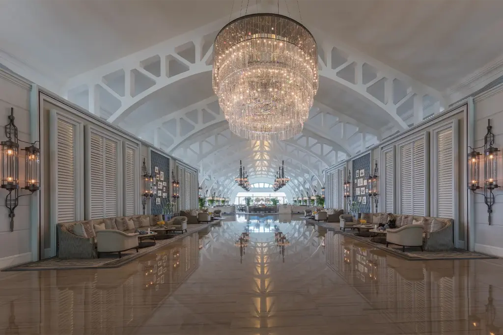 A symmetrical, long-view photograph of a grand luxury hotel lobby featuring vaulted white ceilings, large crystal chandeliers, and plush lounge seating.
