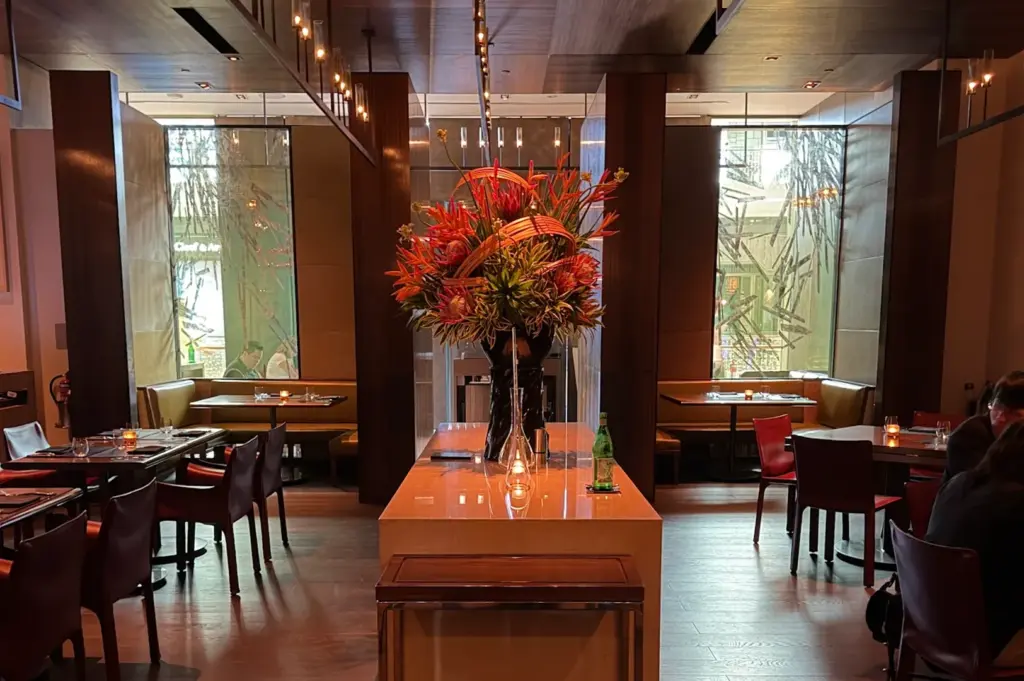 An elegant fine dining room interior at CUT with dark wood pillars, warm ambient lighting, and a large central floral arrangement.