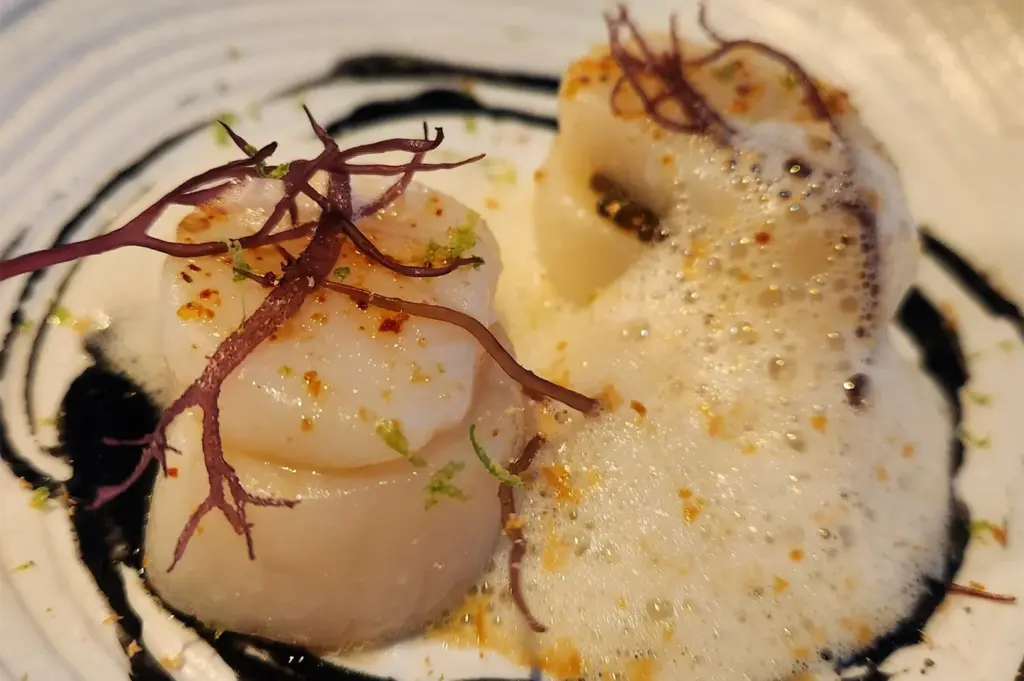 Two scallops garnished with thin red seaweed are plated with a frothy white sauce and vibrant spices on a swirling black and white dish. Luxurious and artistic presentation.