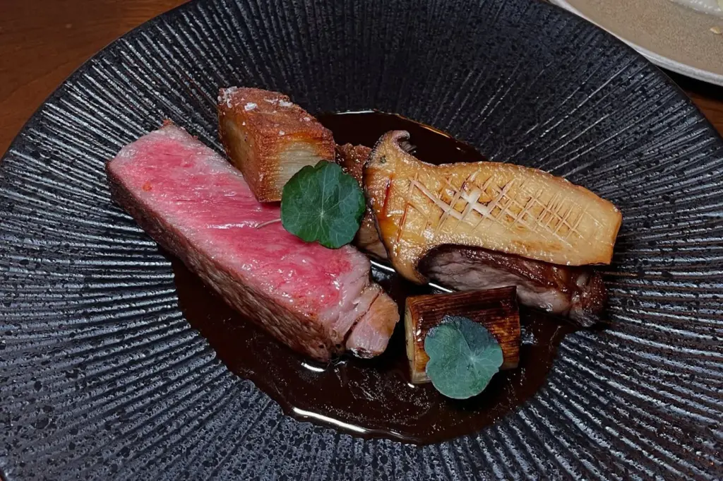 Gourmet dish on a textured black plate featuring a seared pink meat slice, roasted vegetables, mushrooms, garnished with green leaves. Elegant presentation.
