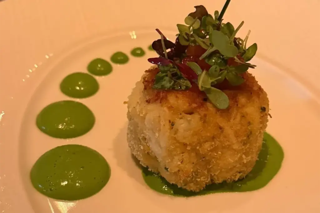 A gourmet jumbo lump crab cake topped with microgreens and served with a decorative swirl of vibrant green pea puree.