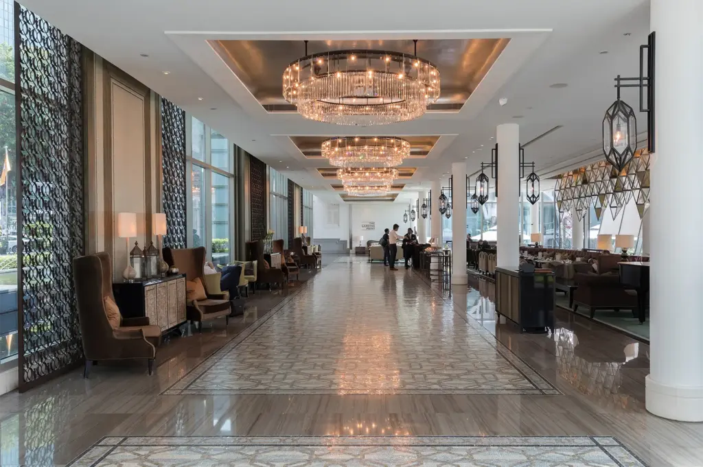 An elegant hotel hallway and lounge area featuring polished marble floors, circular crystal chandeliers, and floor-to-ceiling windows with intricate screen patterns.