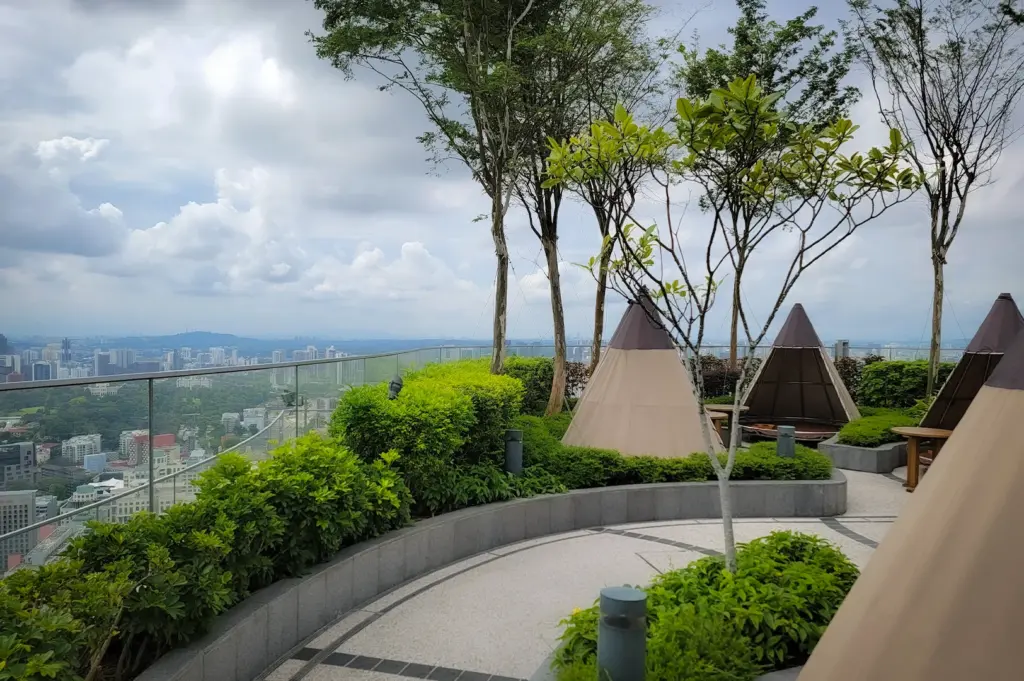 Rooftop garden with teepee-style seating, surrounded by lush greenery and trees. Overlooks a cityscape under a cloud-dotted sky. Calm and inviting.