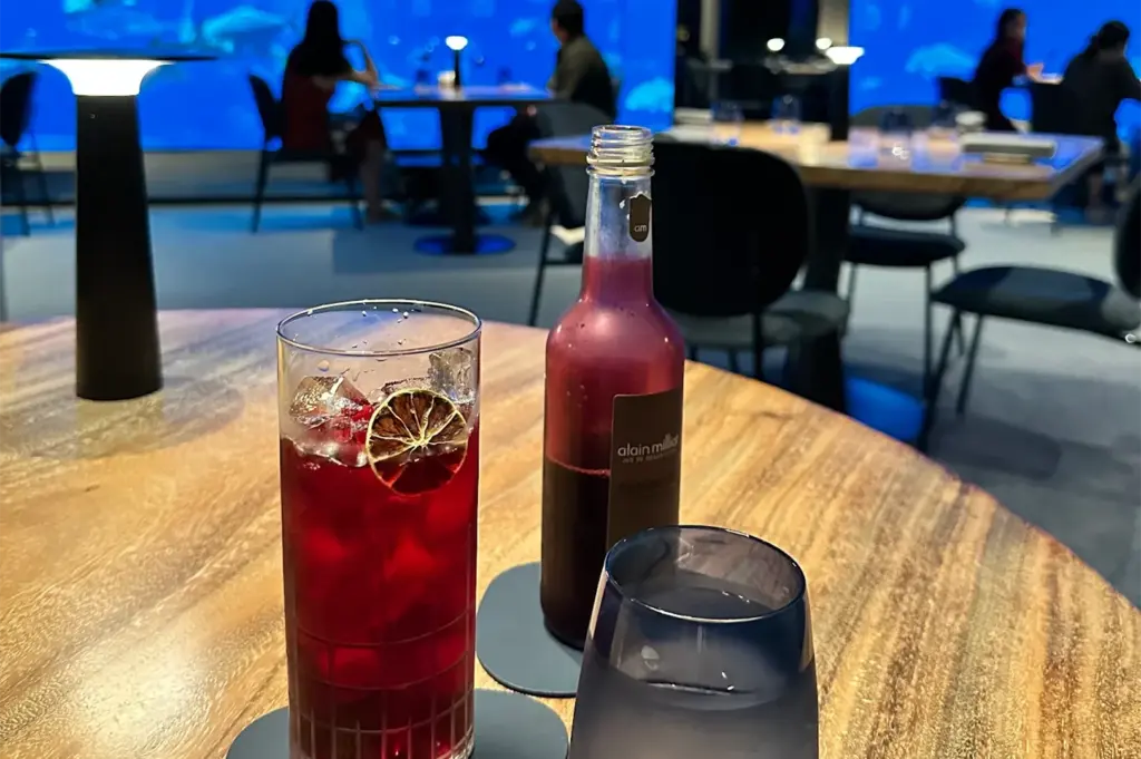 A cozy restaurant scene with dim lighting, showcasing a wooden table holding a red drink with lime, a bottle, and a glass. In the background, silhouettes of people sitting near a large aquarium.