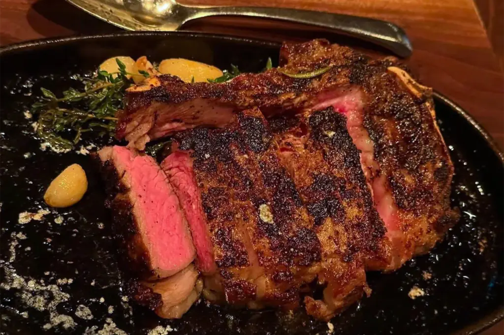 A perfectly seared medium-rare bone-in ribeye steak sliced on a black platter, garnished with roasted garlic and fresh thyme.
