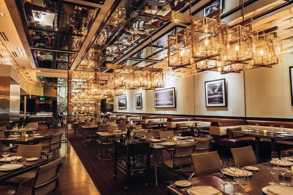 A spacious, high-end restaurant dining room with geometric gold chandeliers, mirrored ceilings, and leather booth seating.