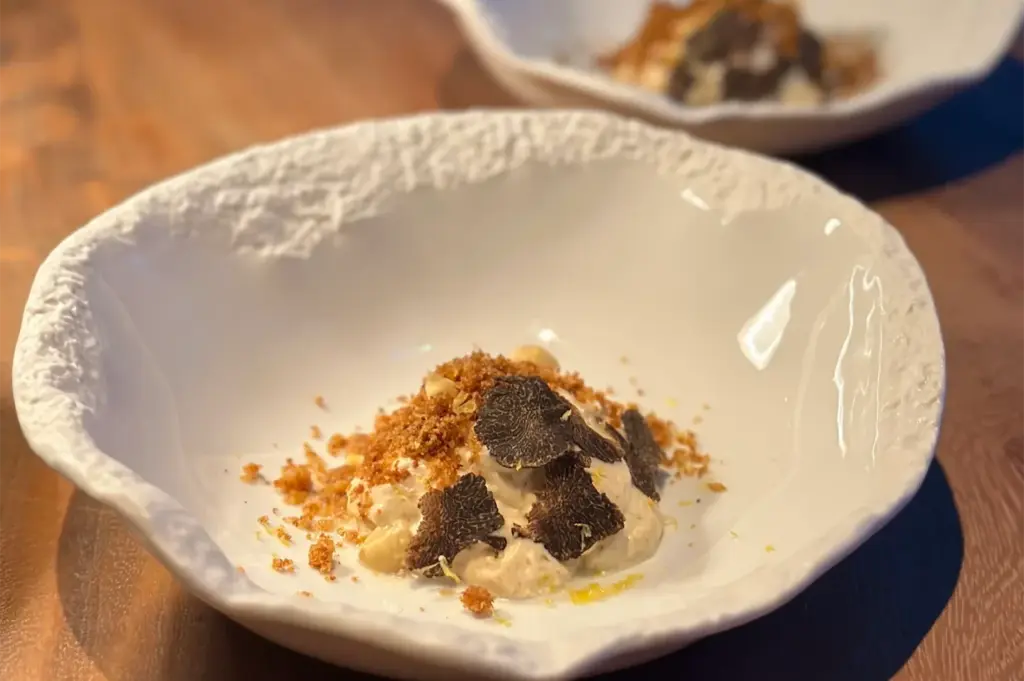 Elegant dish featuring creamy pasta topped with sliced black truffles and a sprinkle of breadcrumbs, served in a textured white bowl on a wooden table.