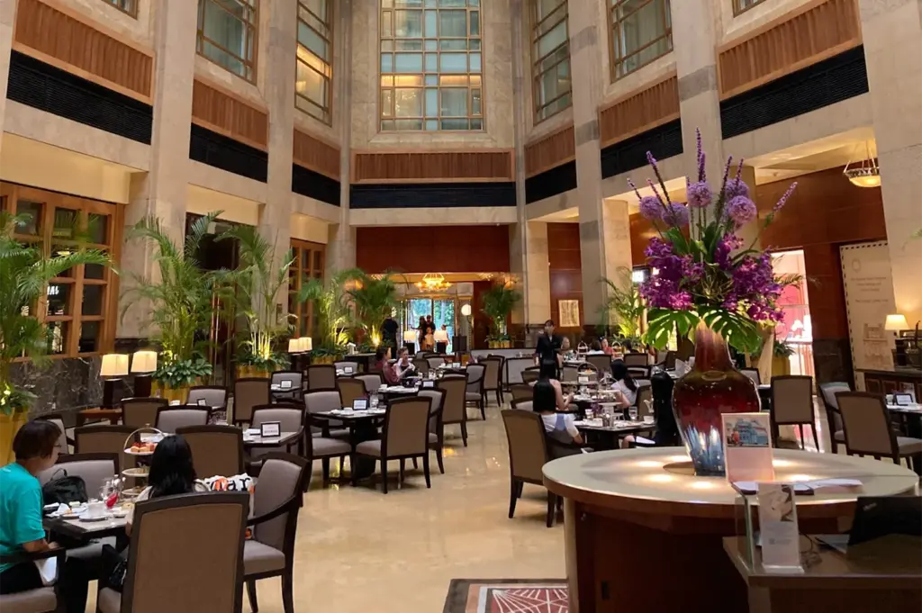 An indoor view of a luxury hotel restaurant with a massive floral centerpiece in the foreground and patrons seated at tables for a formal tea service.