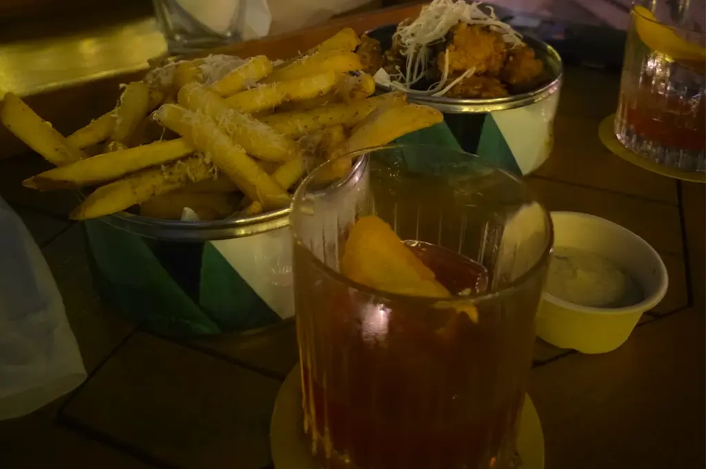 Delicious bar snacks at Mr. Stork, Andaz Singapore, featuring a bucket of golden truffle fries topped with parmesan cheese, crispy fried chicken bites, and a pair of classic cocktails garnished with orange peels on a rustic wooden tabletop.