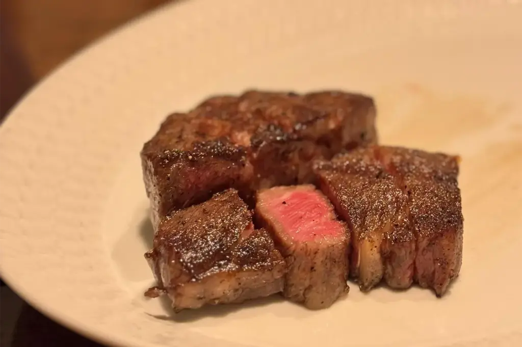 A close-up of a premium thick-cut steak, seared on the outside and medium-rare pink on the inside, served on a white plate.