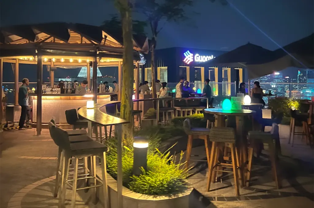 Atmospheric night scene at the Mr. Stork rooftop bar in Singapore, showing patrons enjoying drinks at the circular bar and high tables, with illuminated Suntec and GuocoLand buildings visible in the background.