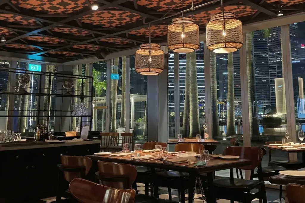 Interior of a sophisticated restaurant at night with large windows overlooking palm trees and the Singapore city skyline.